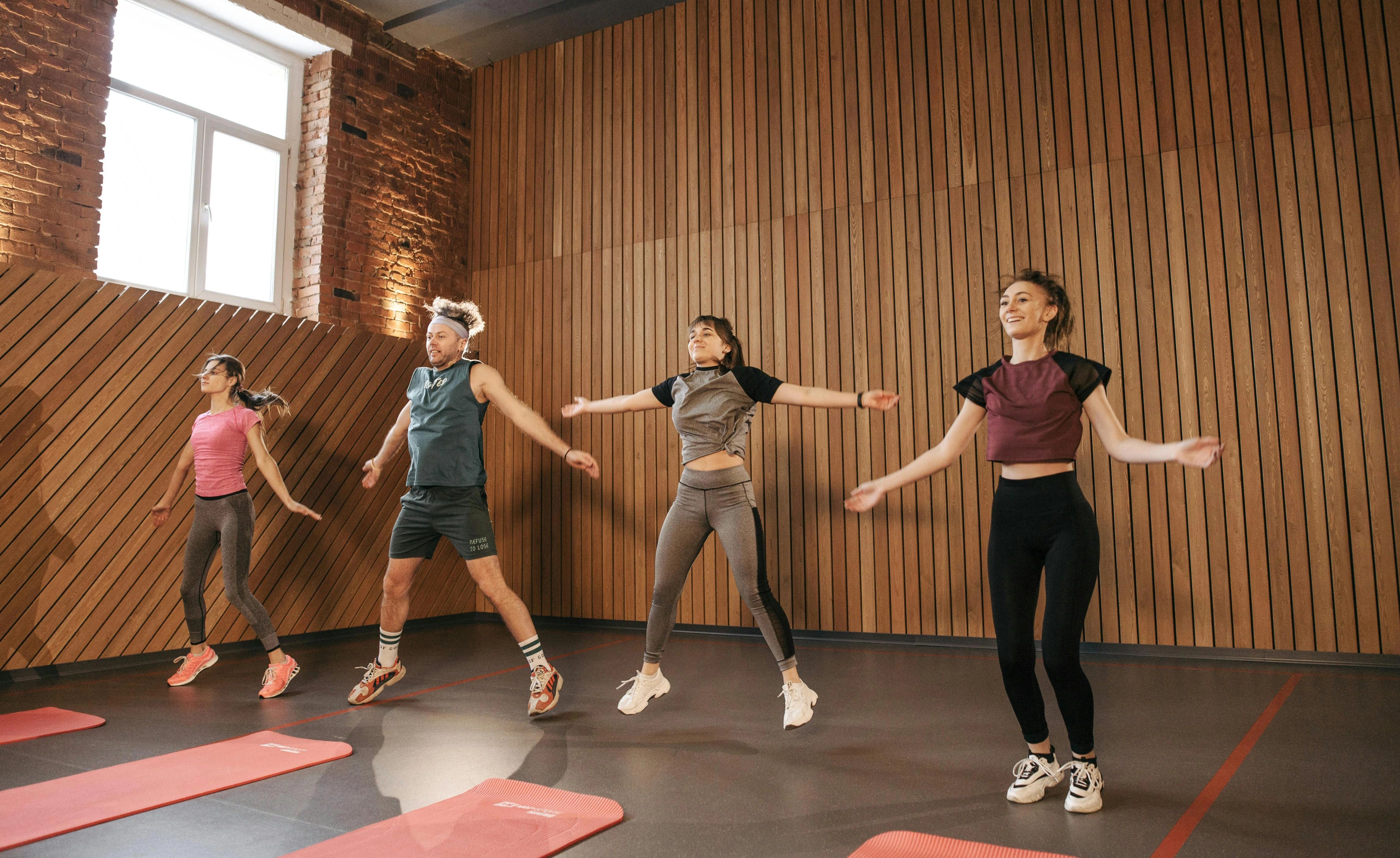 Vier Personen machen gemeinsam Sportübungen in einem Raum mit Holzwänden und großen Fenstern. Sie führen synchron Sprungbewegungen aus, alle tragen sportliche Kleidung. Im Vordergrund liegen rote Trainingsmatten auf dem Boden. Die Szene wirkt aktiv und energiegeladen.