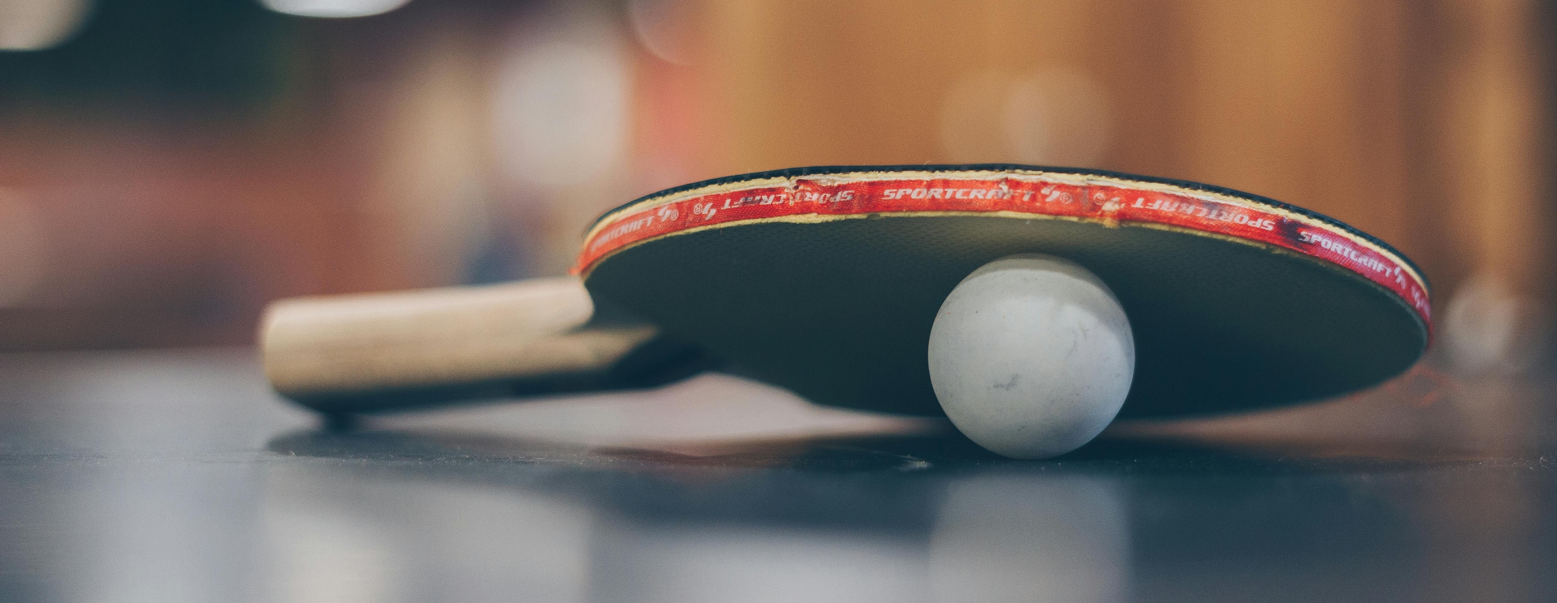 Ein Tischtennisschläger liegt auf einem Tisch, gestützt von einem Tischtennisball. Der Schläger zeigt Abnutzungsspuren, was auf regelmäßigen Gebrauch hinweist. Der Hintergrund ist unscharf und vermittelt eine warme, sportliche Atmosphäre.